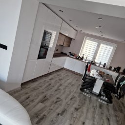 Marci Marcin - Nowoczesna kuchnia z białymi, błyszczącymi frontami szafek, drewnianym blatem i szarymi panelami podłogowymi, widok na stół z czarnymi krzesłami i okno z żaluzjami.