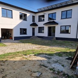 Marci Marcin - Dwupiętrowy dom z jasną elewacją i ciemnymi ramami okiennymi, widok z zewnątrz na podwórze z elementami budowlanymi i garażem z narzędziami.