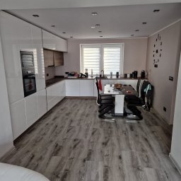 Marci Marcin - Nowoczesna kuchnia z białymi, lakierowanymi szafkami, drewnianymi akcentami, szarą podłogą imitującą drewno i designerskim zegarem ściennym.