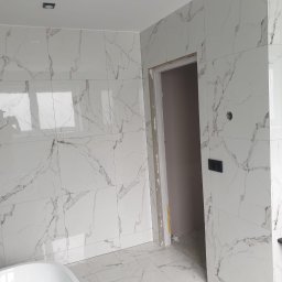 Marci Marcin - Świeżo wykafelkowana łazienka z białymi płytkami imitującymi marmur, widoczny fragment wanny i czarna szafka pod umywalkę, surowa ościeżnica drzwi.