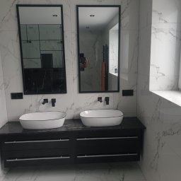 Marci Marcin - Nowoczesna łazienka w Radomsku z dwoma umywalkami nablatowymi, czarnym blatem i szafkami oraz dużymi lustrami w czarnych ramach na tle białych płytek imitujących marmur.