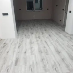 Jasne panele podłogowe imitujące drewno, ułożone w pustym pomieszczeniu z przygotowanymi gniazdkami elektrycznymi i oknem w tle.