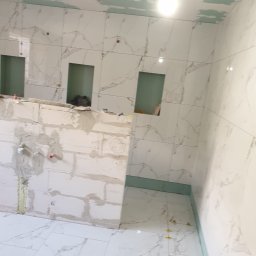 Marci Marcin - Wnętrze łazienki w trakcie prac: białe płytki imitujące marmur na ścianach z wnękami, surowa ściana z białych bloczków, częściowo położona podłoga z płytek, widoczne krzyżyki dystansowe, sufit...