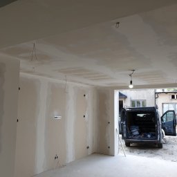 Marci Marcin - Surowe ściany wewnątrz remontowanego pomieszczenia z widoczną instalacją elektryczną i samochodem dostawczym zaparkowanym na zewnątrz.