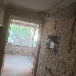 Marci Marcin - Wnętrze w trakcie prac przygotowawczych: odsłonięta cegła na ścianach, widoczne przewody elektryczne, okno z widokiem na zieleń, materiały budowlane zgromadzone na parapecie.