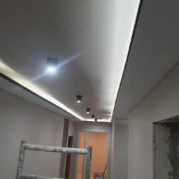 Marci Marcin - Sufit z zamontowanym oświetleniem LED wzdłuż krawędzi, widoczne czarne reflektory punktowe, na pierwszym planie drabina malarska z resztkami farby, w tle widoczne drzwi do innego pomieszczenia.
