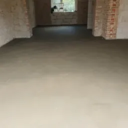 Wylany, świeży podkład podłogowy w pomieszczeniu z odkrytą cegłą na fragmentach ścian i widoczną konstrukcją sufitu.