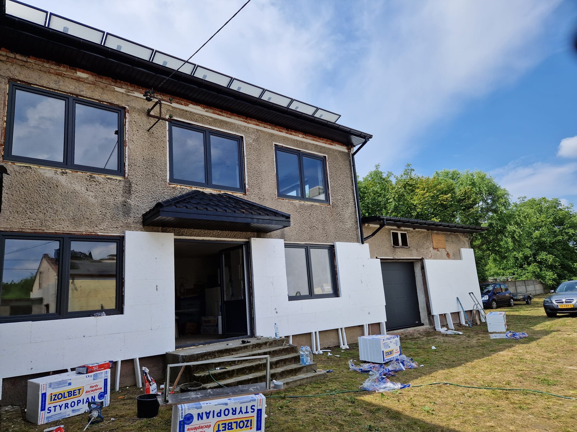 Dom w trakcie ocieplania styropianem, widoczne płyty izolacyjne Izolet Styropian, okna z czarnymi ramami, panele solarne na dachu, teren wokół domu w trakcie prac.