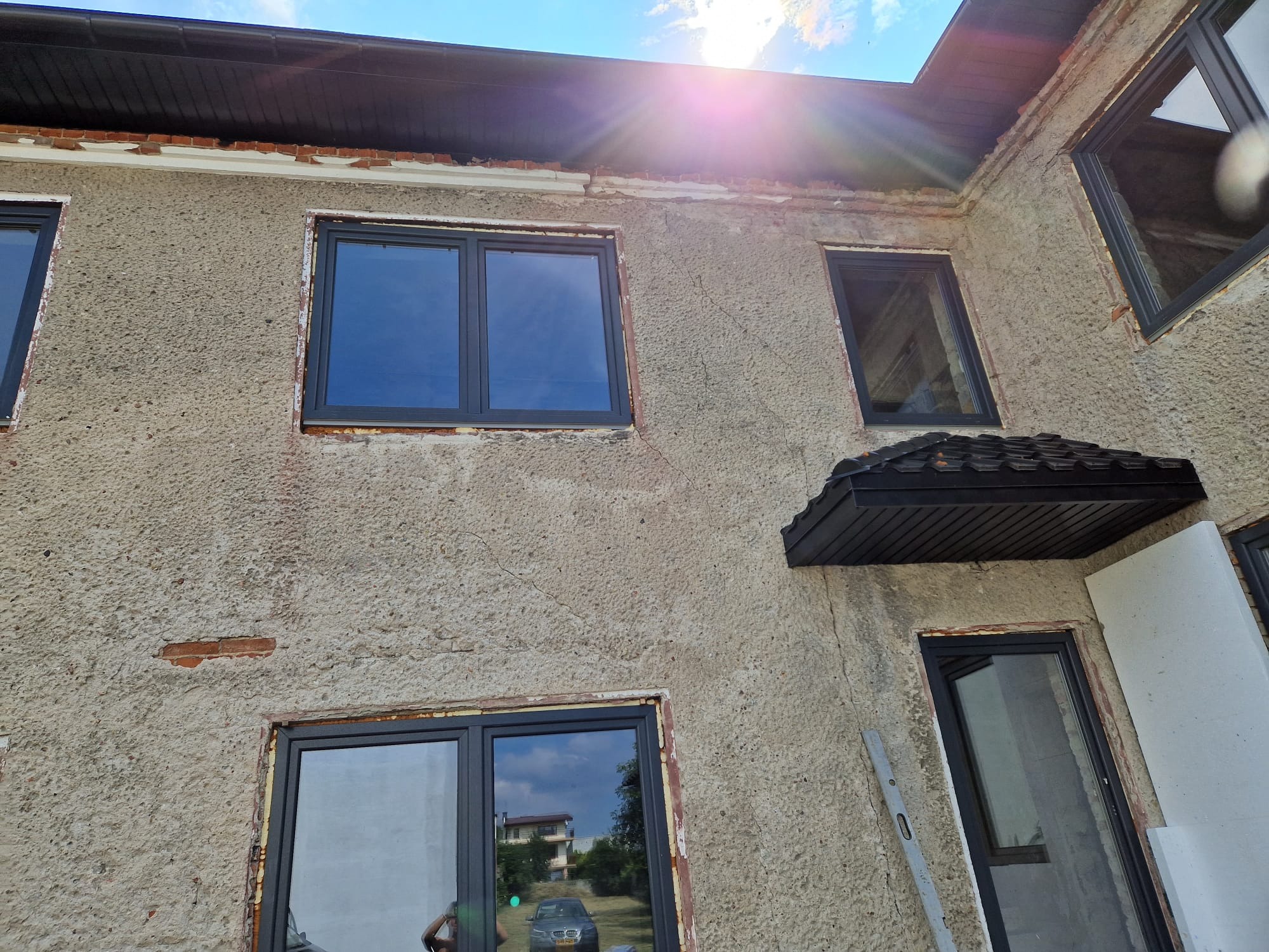 Elewacja budynku z nowymi, grafitowymi oknami przed ociepleniem, widoczne fragmenty starej, uszkodzonej elewacji i brakujące cegły, drabina oparta o ścianę.