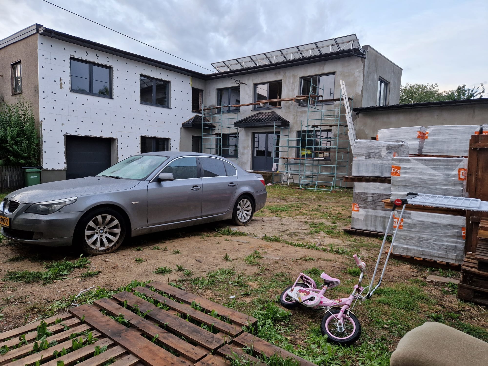 Dom w trakcie ocieplania, widoczne rusztowania, częściowo ocieplona elewacja styropianem, na dachu panele słoneczne, obok zaparkowany szary samochód, na pierwszym planie palety i różowy rowerek...