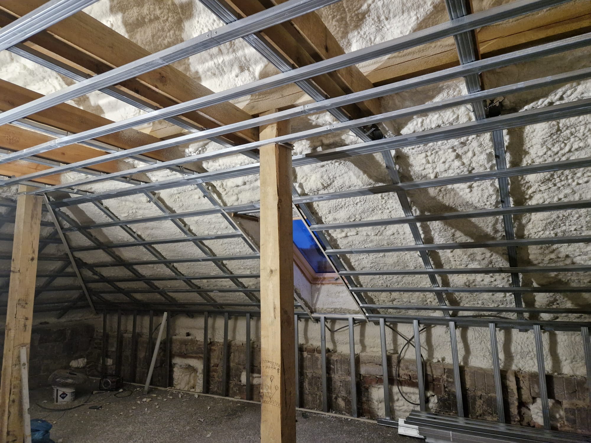 Poddasze w trakcie ocieplania pianką PUR, widoczne krokwie, metalowy stelaż pod płyty gipsowo-kartonowe i częściowo nałożona pianka, z niebieskim oknem dachowym w tle i drewnianym słupem...