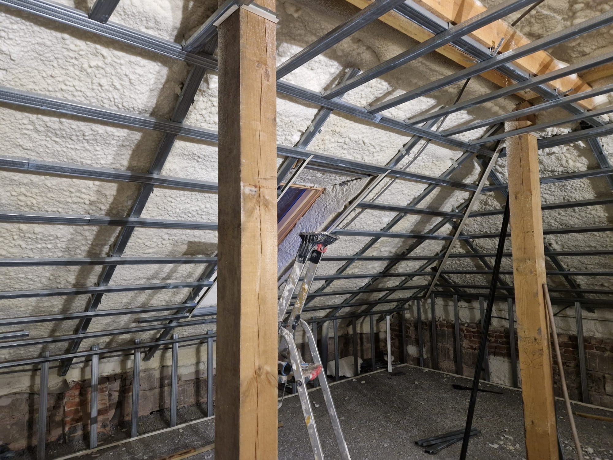 Poddasze w trakcie prac: ocieplenie pianką PUR na skośnym dachu, widoczne metalowe profile konstrukcyjne, drewniane słupy podtrzymujące, drabina i odsłonięta cegła na ścianach