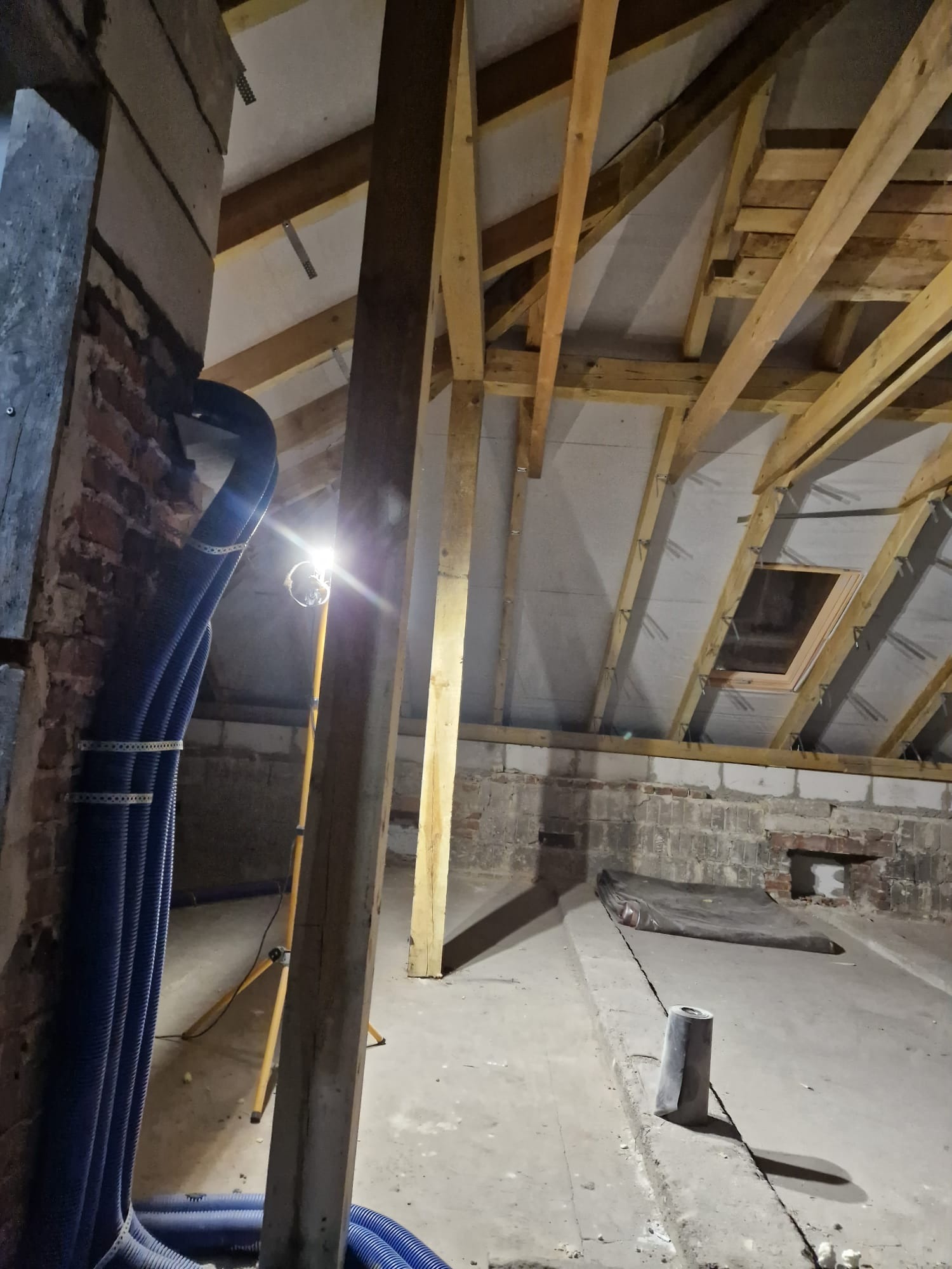 Poddasze w trakcie remontu, widoczne drewniane krokwie, izolacja, niebieskie rury wentylacyjne mocowane do ściany z cegły, oświetlenie robocze