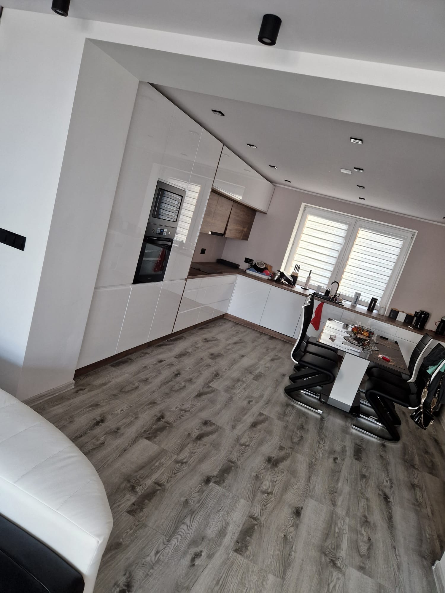 Nowoczesna kuchnia z białymi, błyszczącymi frontami szafek, drewnianym blatem i szarymi panelami podłogowymi, widok na stół z czarnymi krzesłami i okno z żaluzjami.