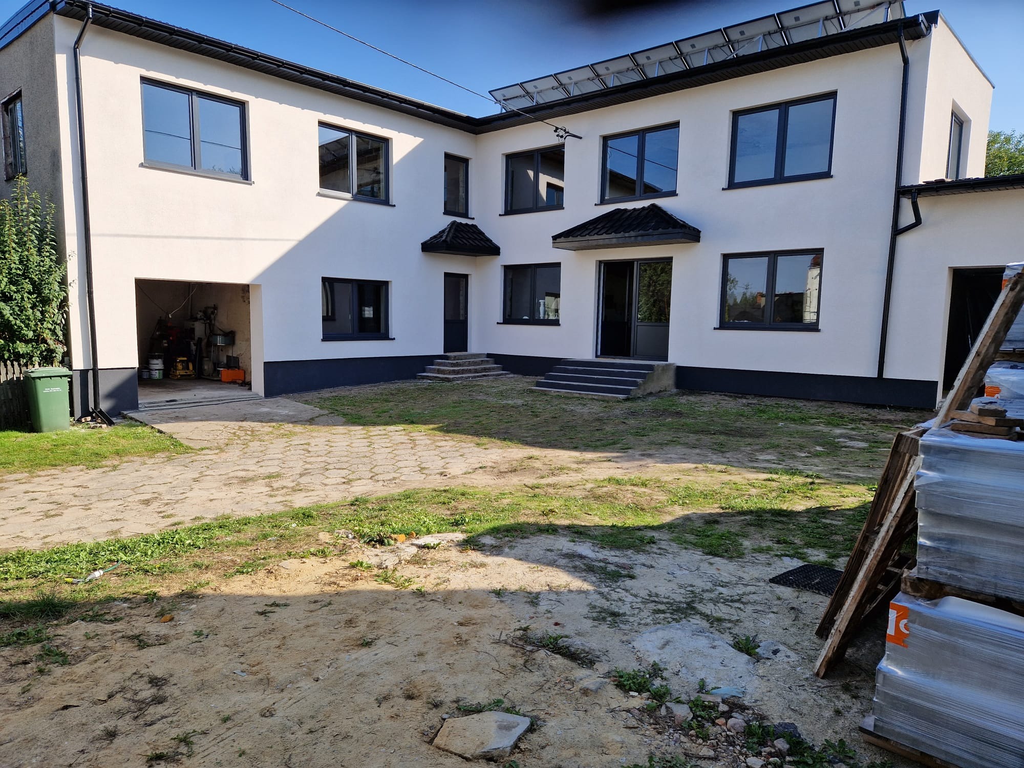 Dwupiętrowy dom z jasną elewacją i ciemnymi ramami okiennymi, widok z zewnątrz na podwórze z elementami budowlanymi i garażem z narzędziami.