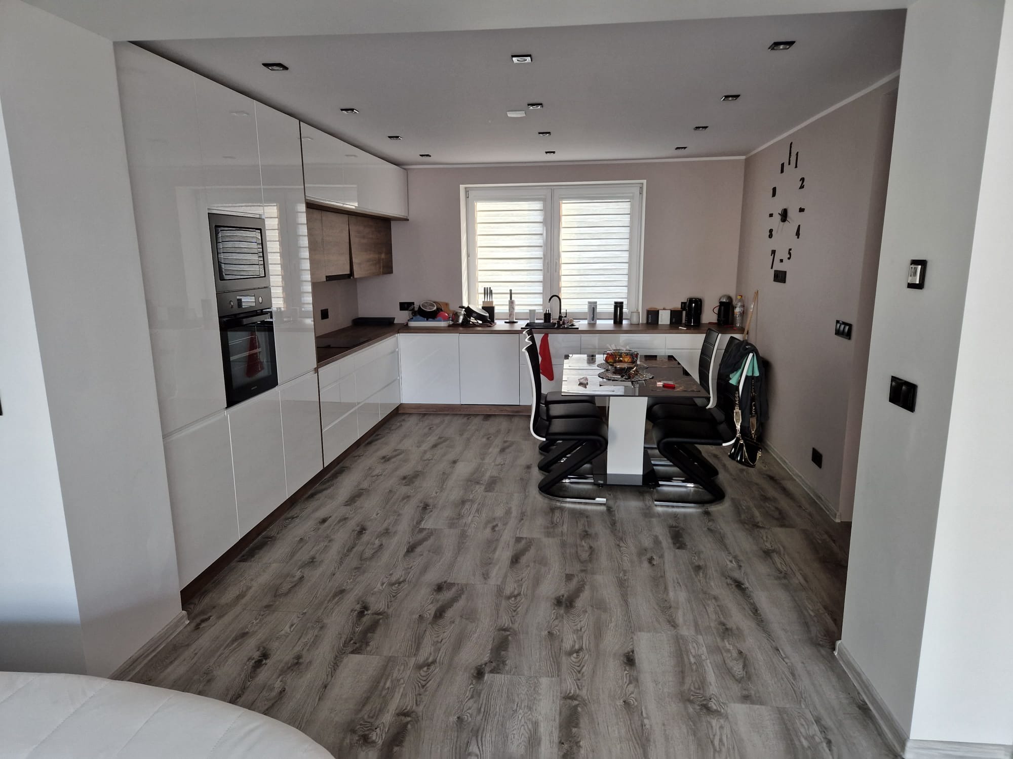 Nowoczesna kuchnia z białymi, lakierowanymi szafkami, drewnianymi akcentami, szarą podłogą imitującą drewno i designerskim zegarem ściennym.