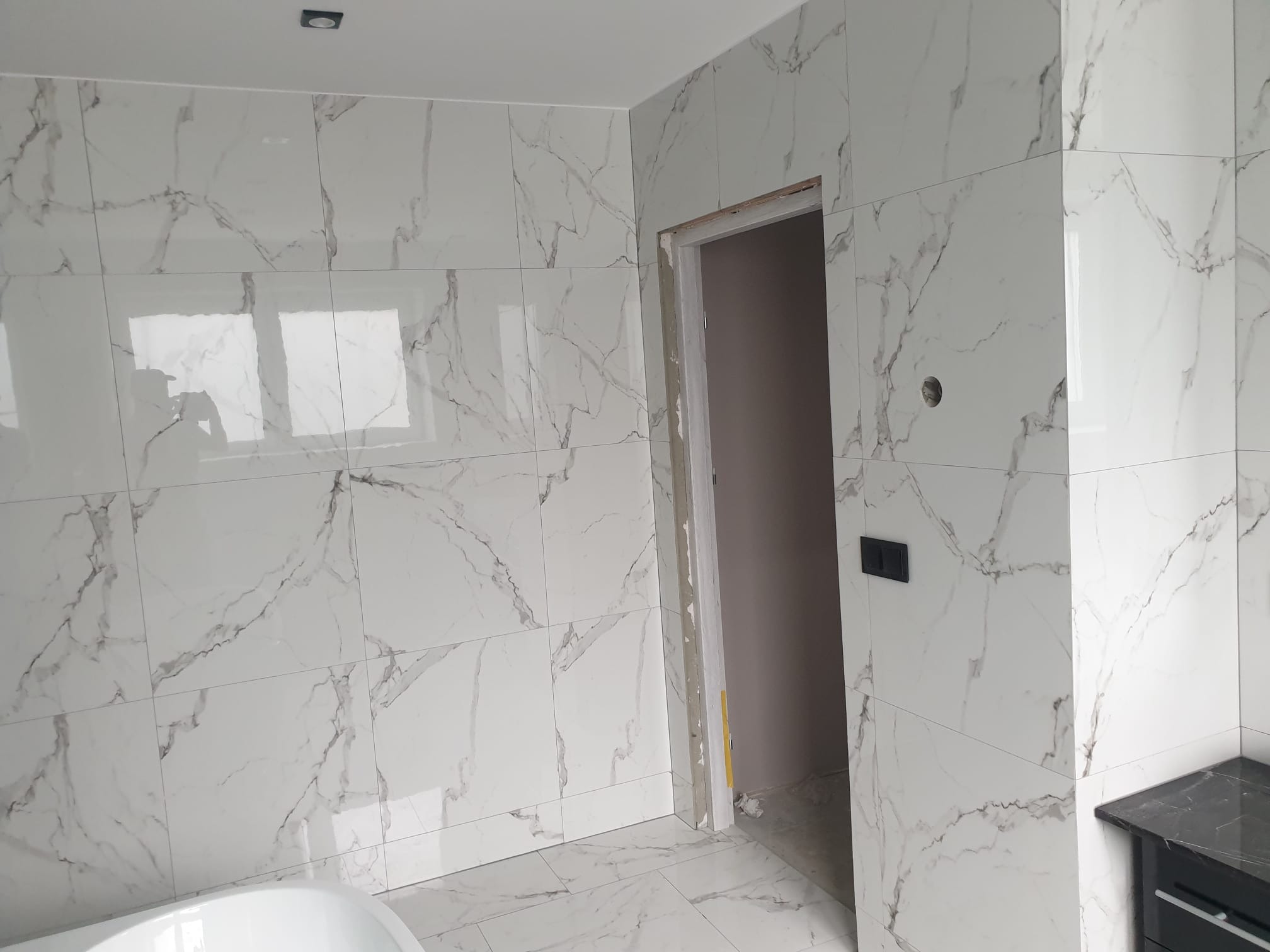 Świeżo wykafelkowana łazienka z białymi płytkami imitującymi marmur, widoczny fragment wanny i czarna szafka pod umywalkę, surowa ościeżnica drzwi.