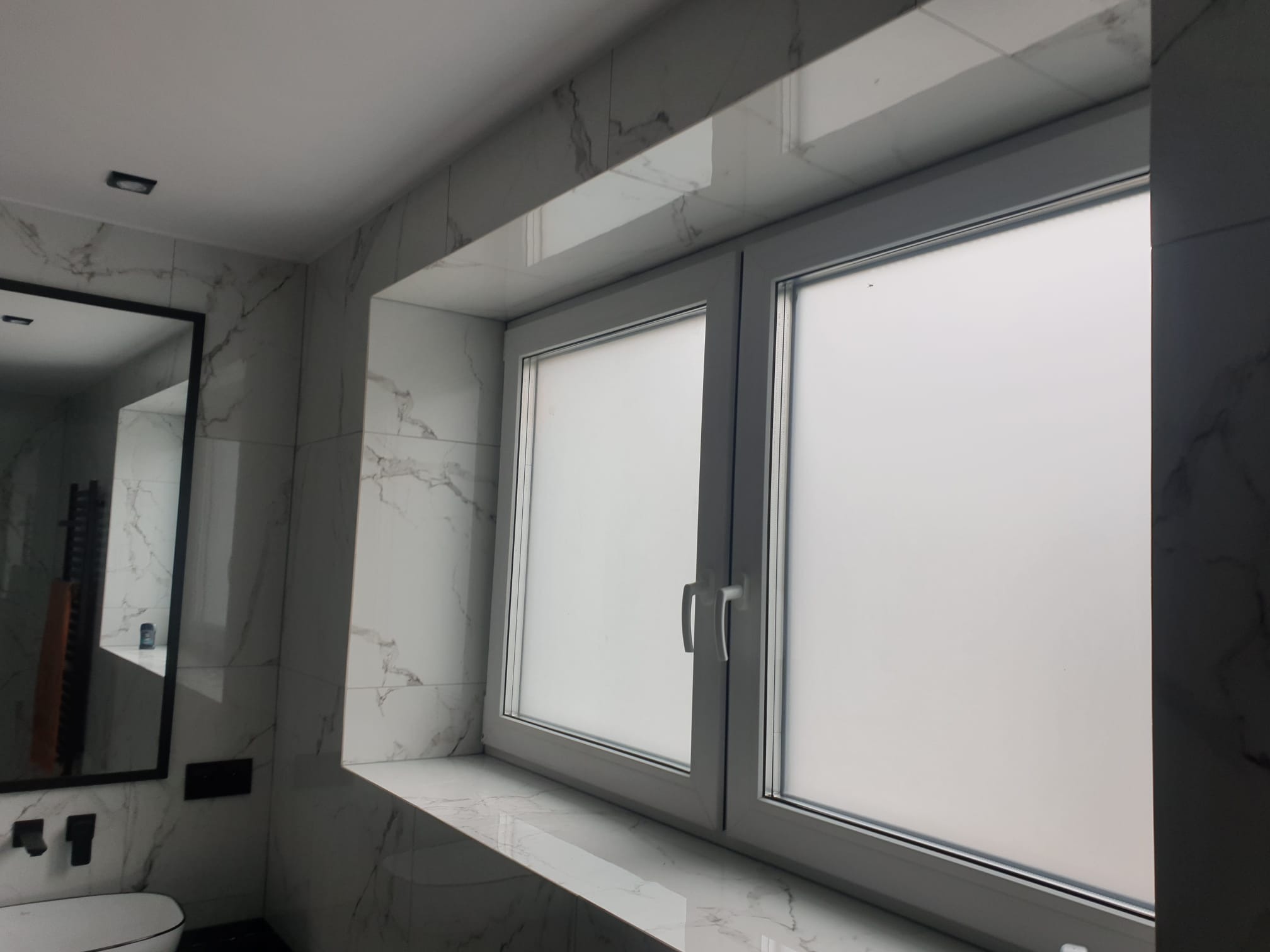 Nowoczesna łazienka z marmurowymi płytkami, białymi oknami z matową szybą i czarnym lustrem wiszącym nad toaletą.