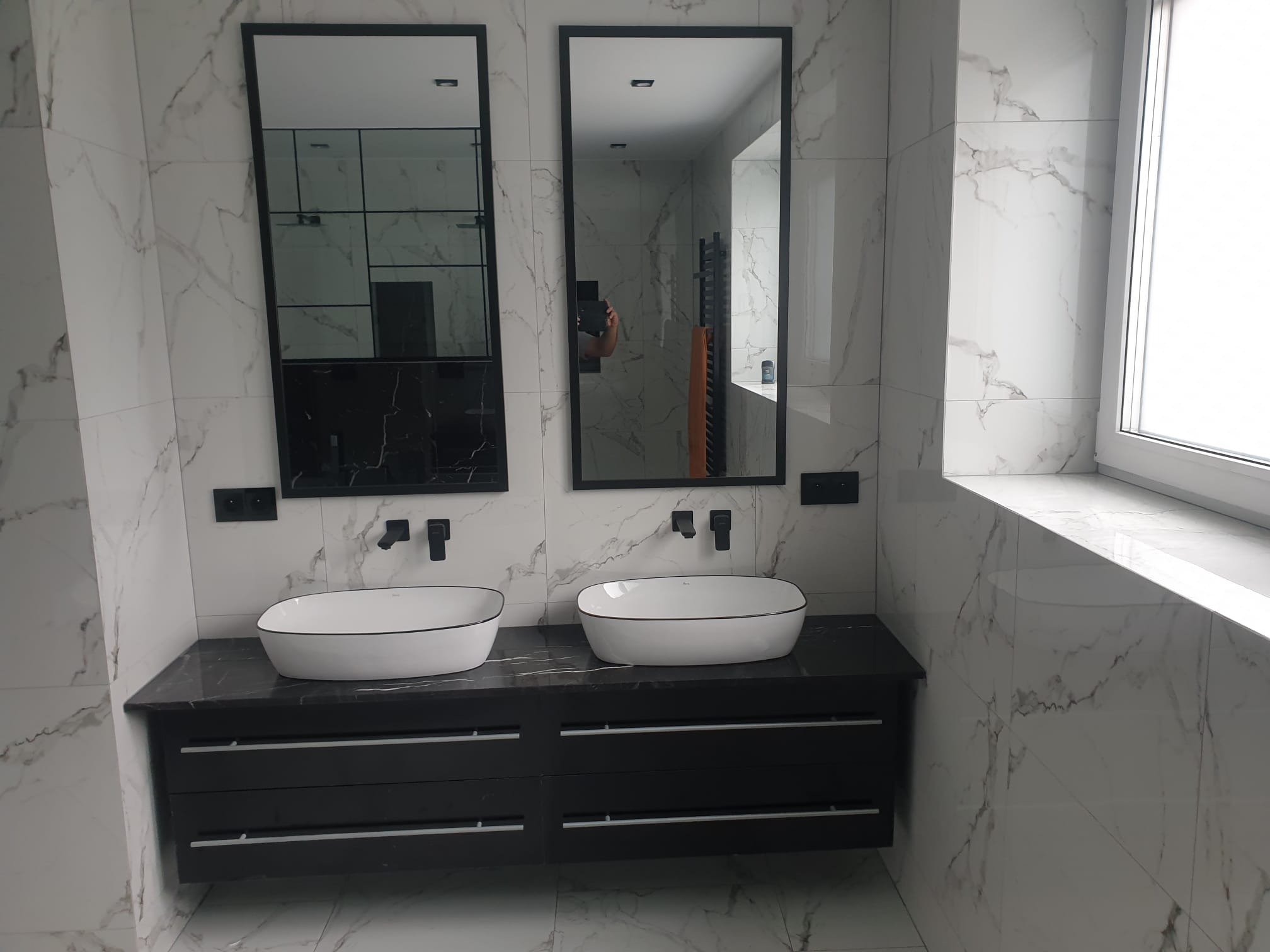 Nowoczesna łazienka w Radomsku z dwoma umywalkami nablatowymi, czarnym blatem i szafkami oraz dużymi lustrami w czarnych ramach na tle białych płytek imitujących marmur.