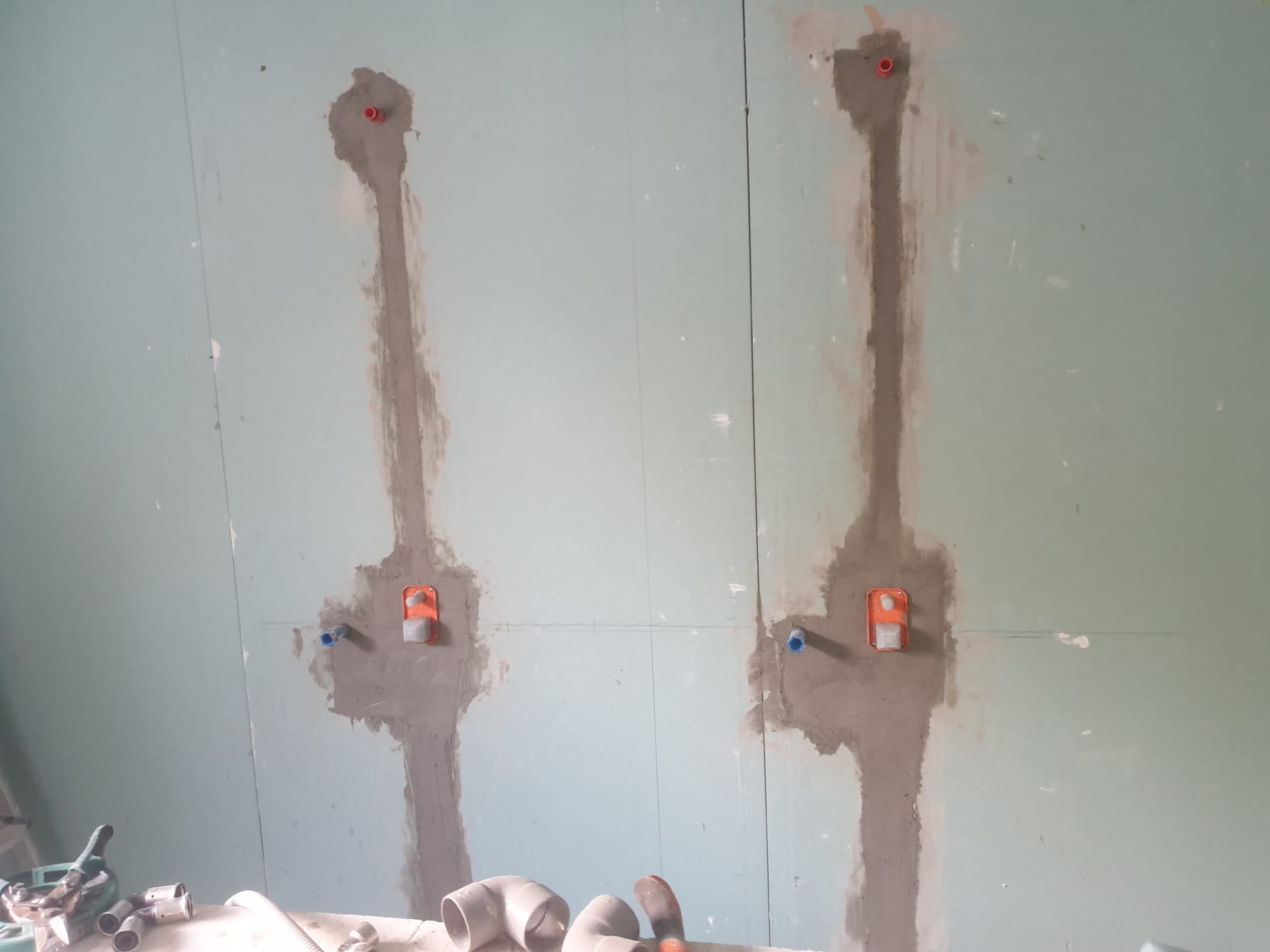 Przygotowanie ściany z płyt gipsowo-kartonowych do montażu armatury łazienkowej, widoczne kable i rury w szpachli oraz narzędzia na dole kadru.