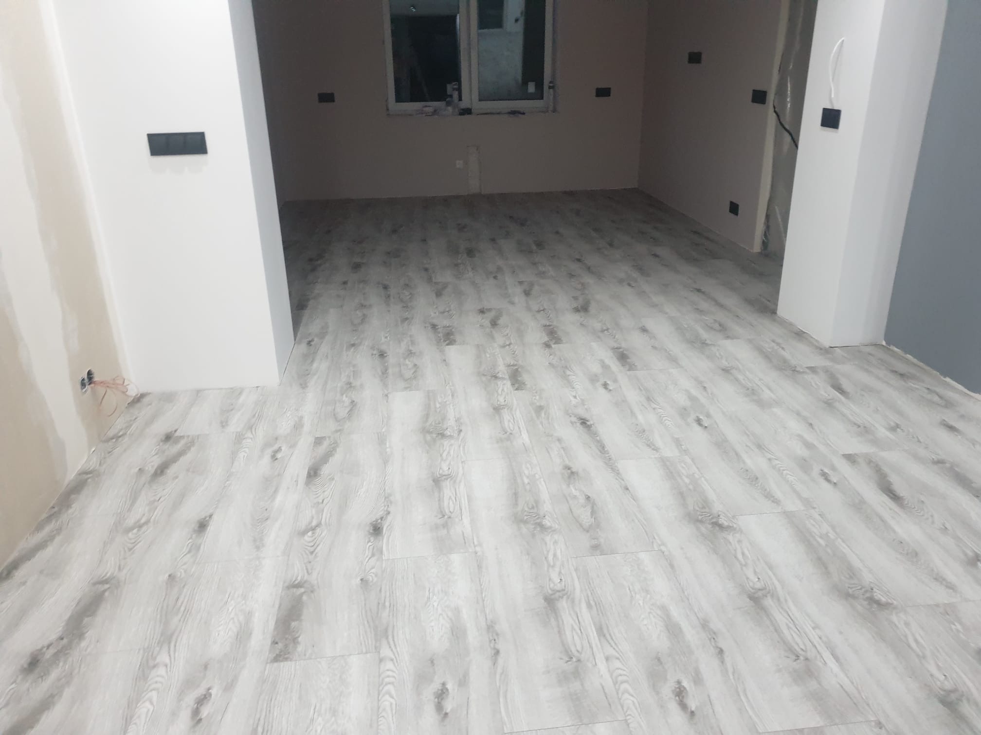 Jasne panele podłogowe imitujące drewno, ułożone w pustym pomieszczeniu z przygotowanymi gniazdkami elektrycznymi i oknem w tle.