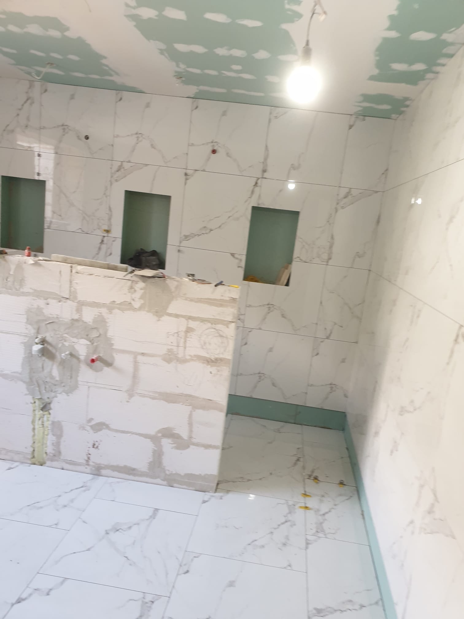 Wnętrze łazienki w trakcie prac: białe płytki imitujące marmur na ścianach z wnękami, surowa ściana z białych bloczków, częściowo położona podłoga z płytek, widoczne krzyżyki dystansowe, sufit...