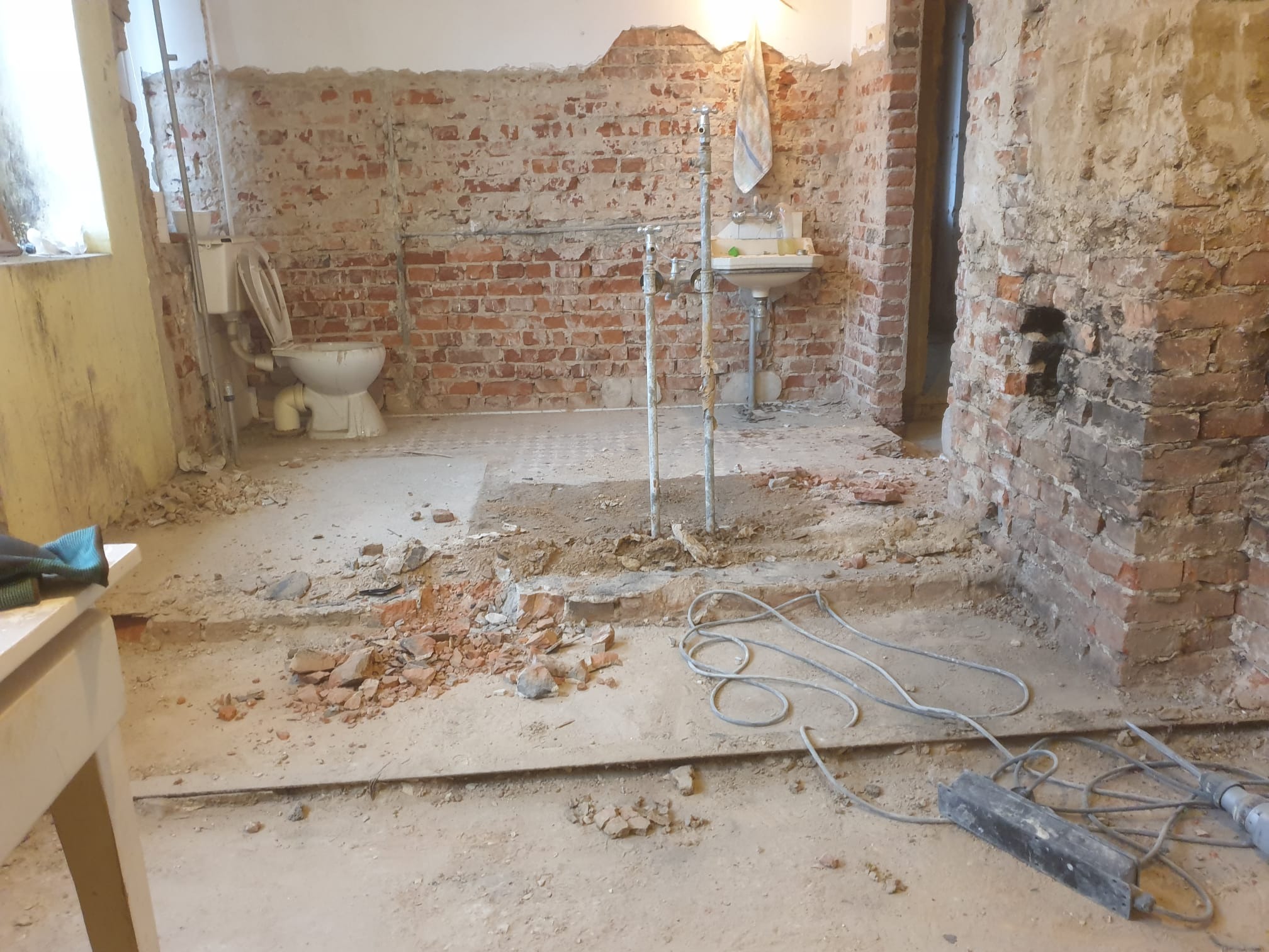 Demontaż starej łazienki: odsłonięta cegła, zdemontowana podłoga, wisząca umywalka i toaleta w trakcie prac rozbiórkowych.