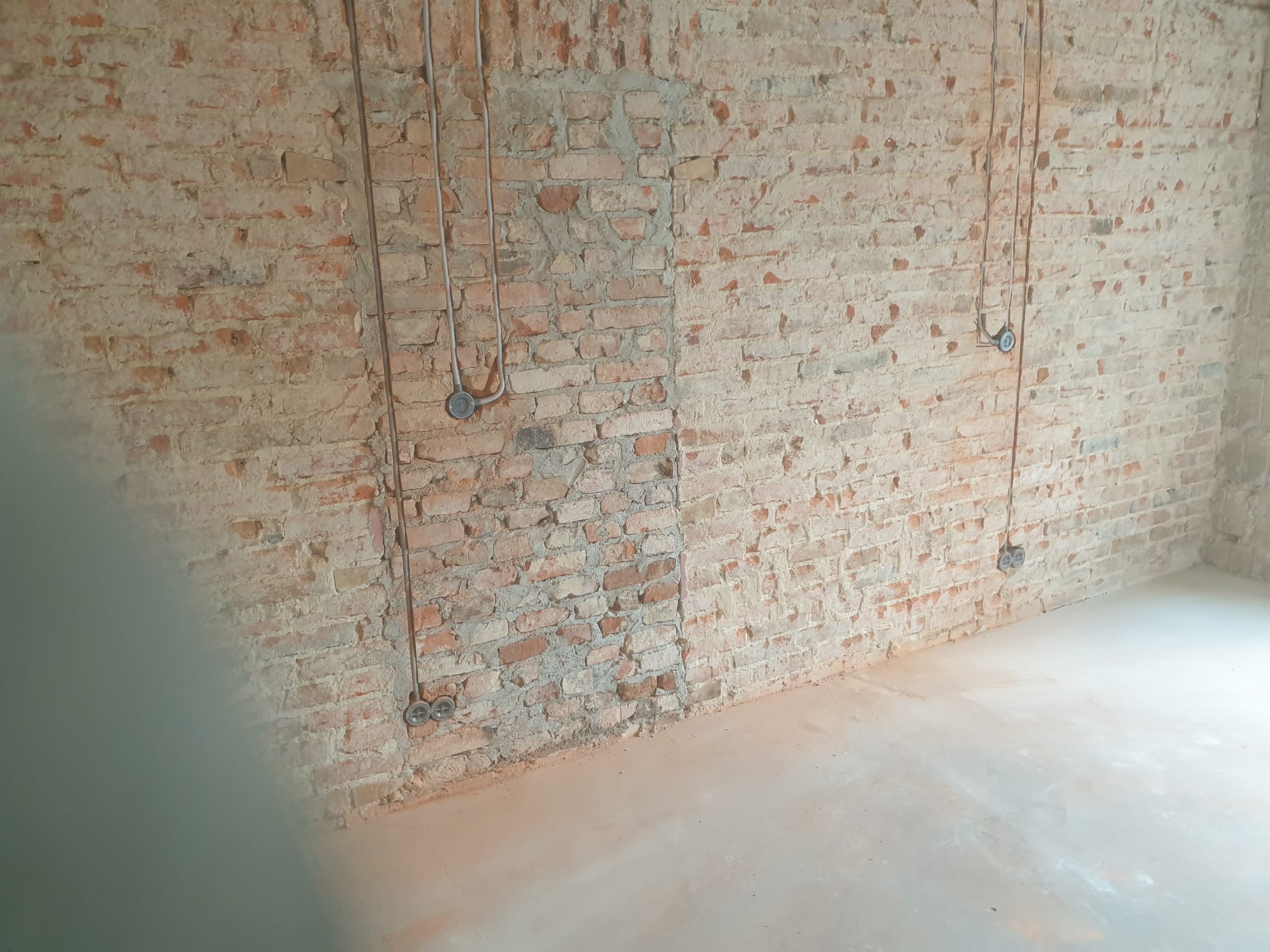 Surowa ściana z cegły z widocznymi przewodami elektrycznymi i puszkami instalacyjnymi, przygotowana do dalszych prac wykończeniowych.