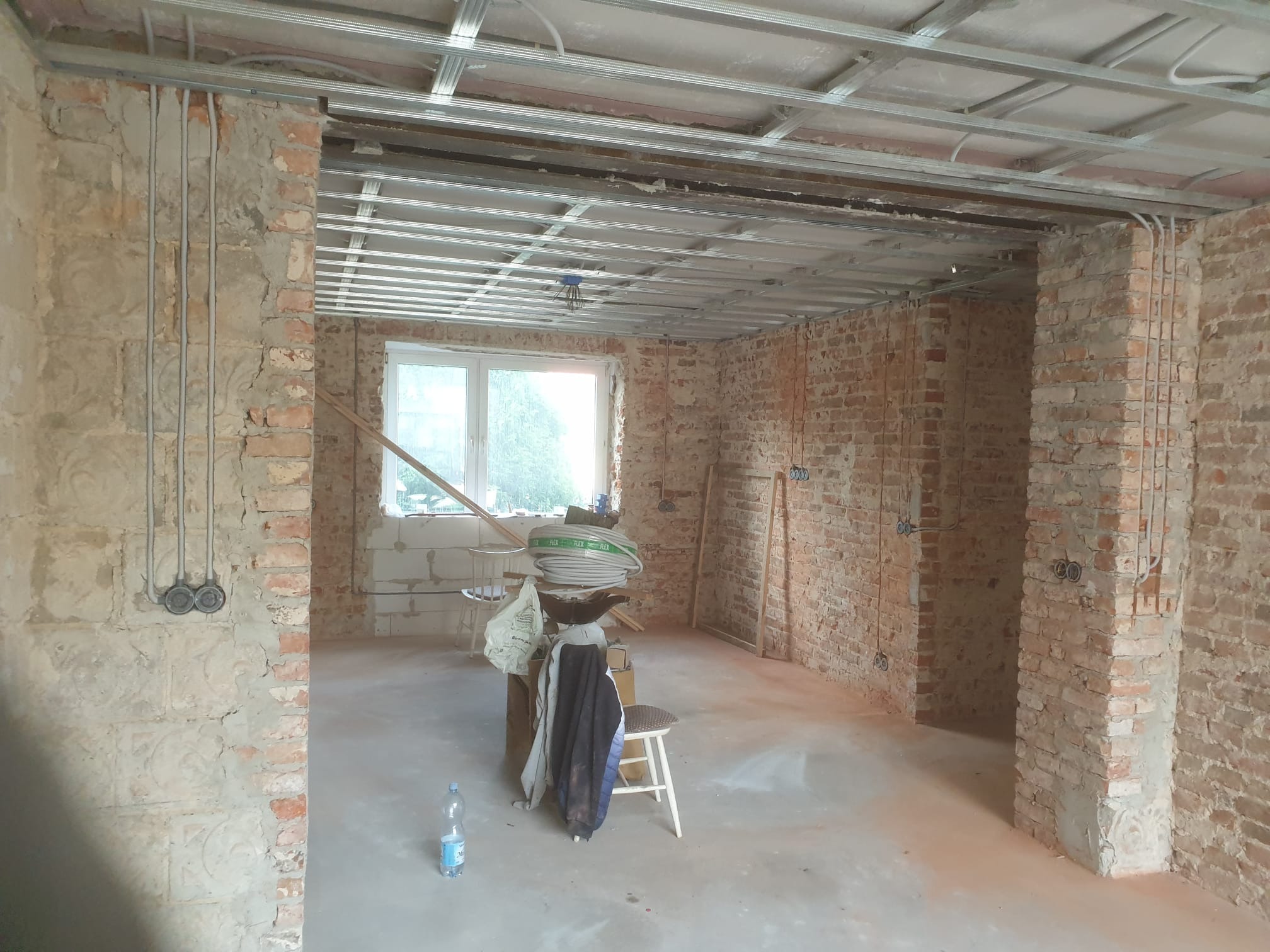 Surowe wnętrze w trakcie prac: odsłonięta cegła, instalacja elektryczna w ścianach, metalowa konstrukcja sufitu podwieszanego, okno w tle, butelka wody na podłodze, krzesło i zwój kabli...