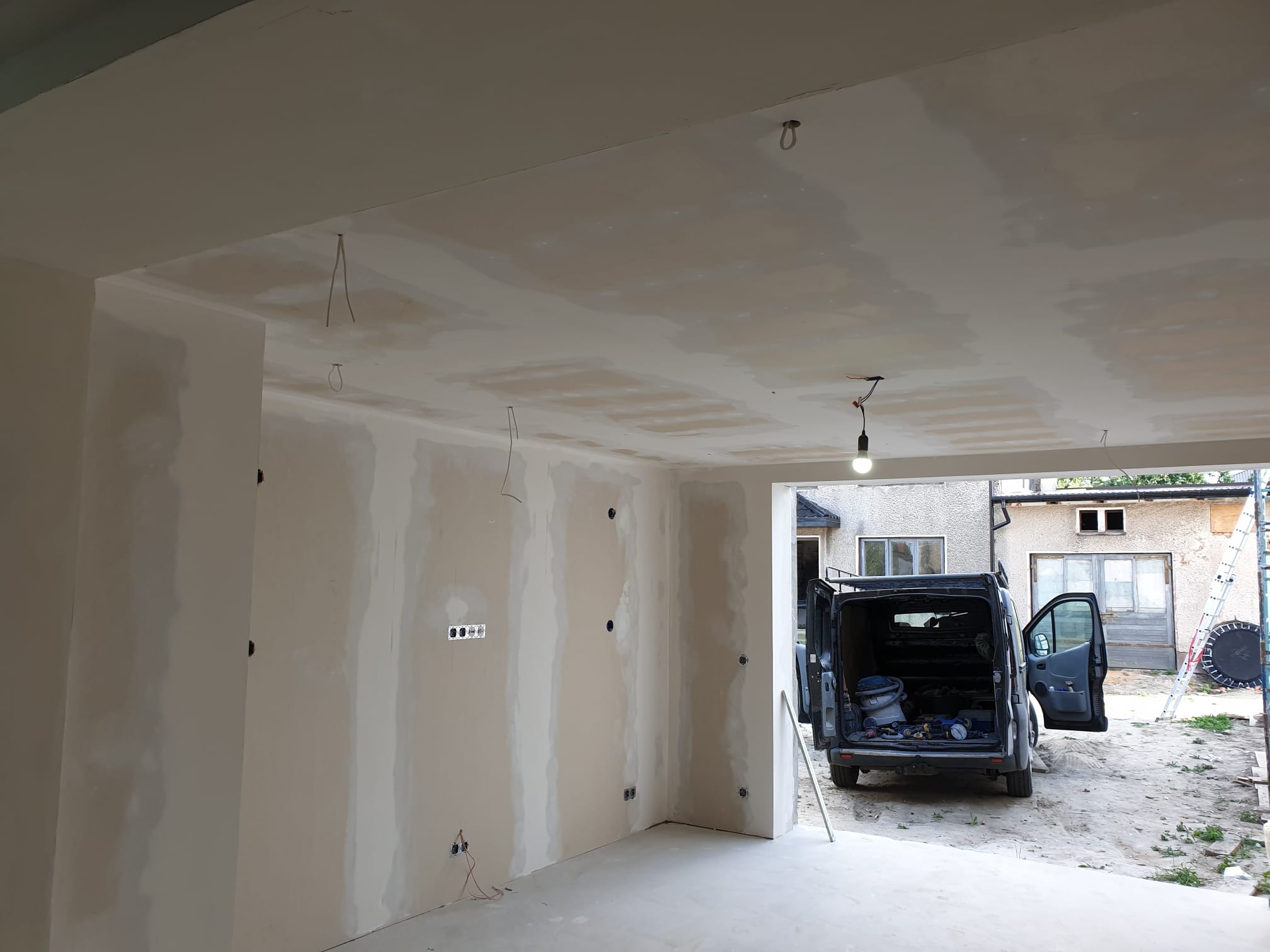 Surowe ściany wewnątrz remontowanego pomieszczenia z widoczną instalacją elektryczną i samochodem dostawczym zaparkowanym na zewnątrz.