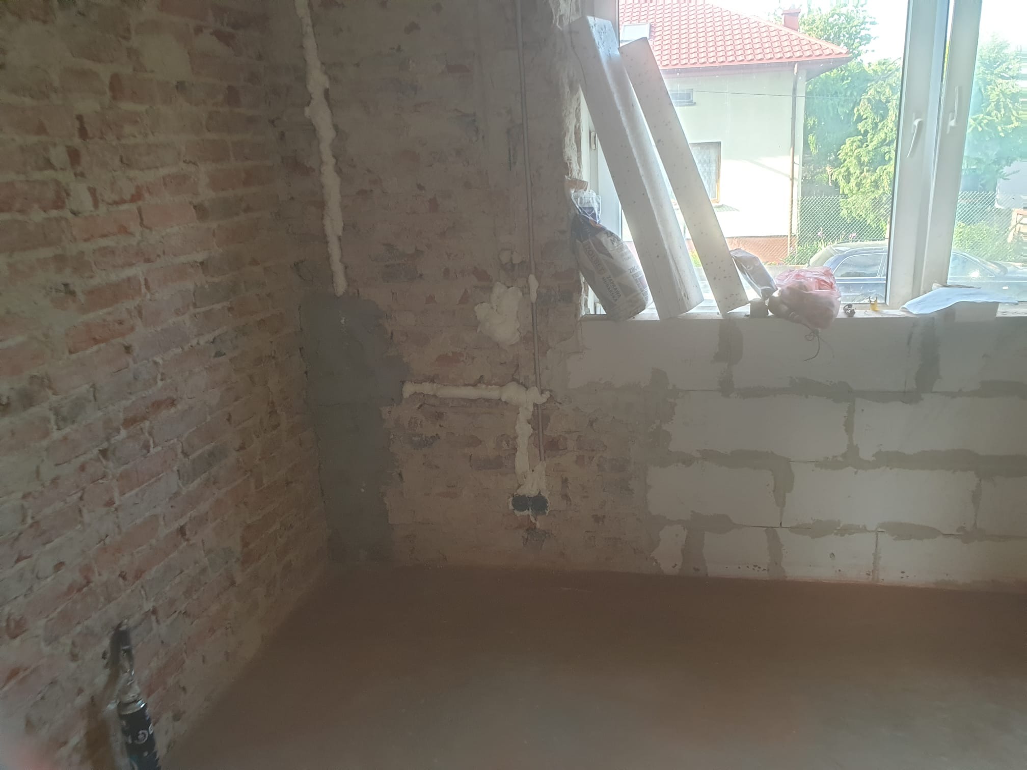 Wnętrze w trakcie remontu: fragment ściany z cegły, instalacja elektryczna zabezpieczona pianką montażową, okno z widokiem na dom i drzewa, torby z materiałami budowlanymi.