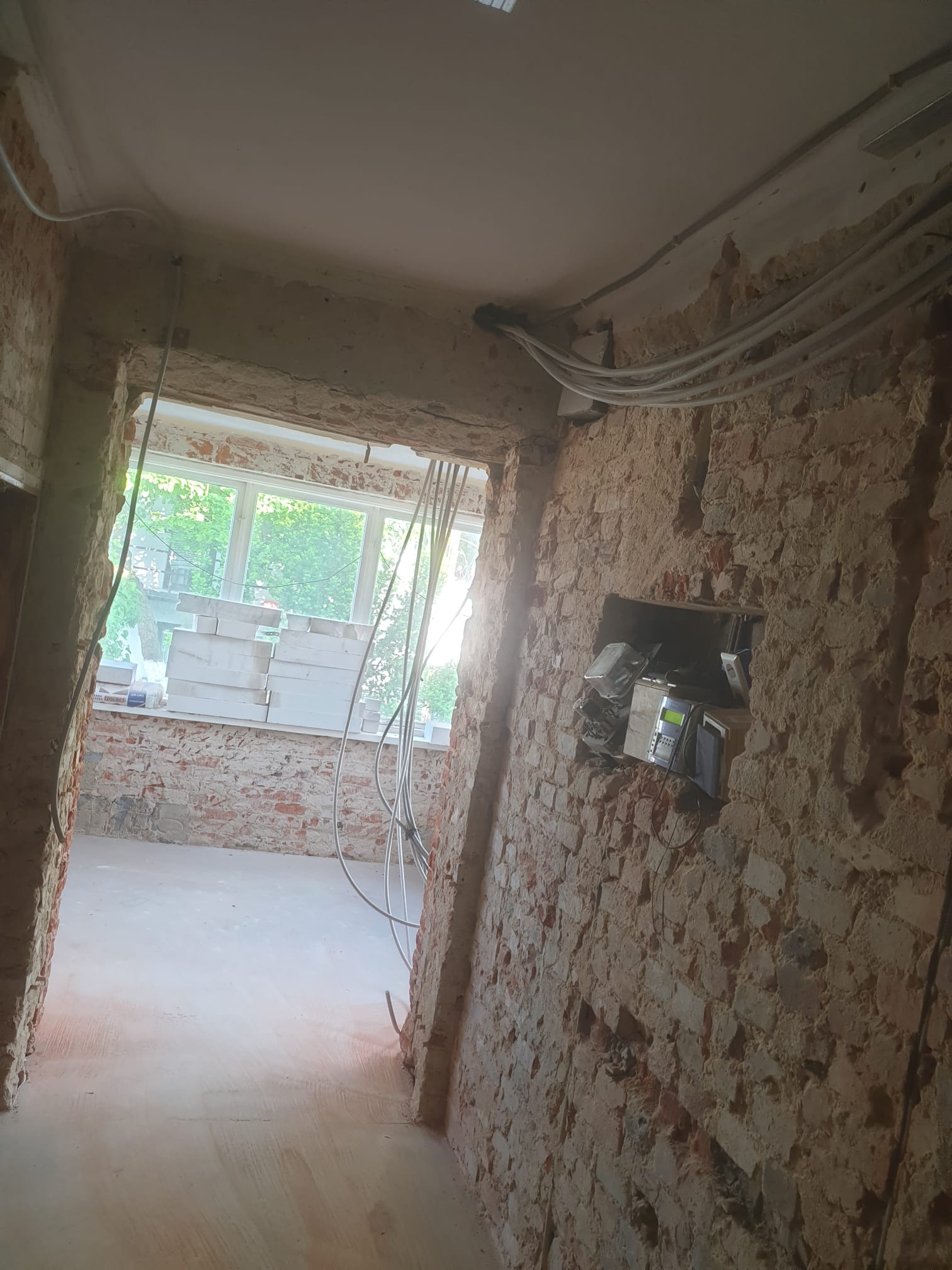 Wnętrze w trakcie prac przygotowawczych: odsłonięta cegła na ścianach, widoczne przewody elektryczne, okno z widokiem na zieleń, materiały budowlane zgromadzone na parapecie.