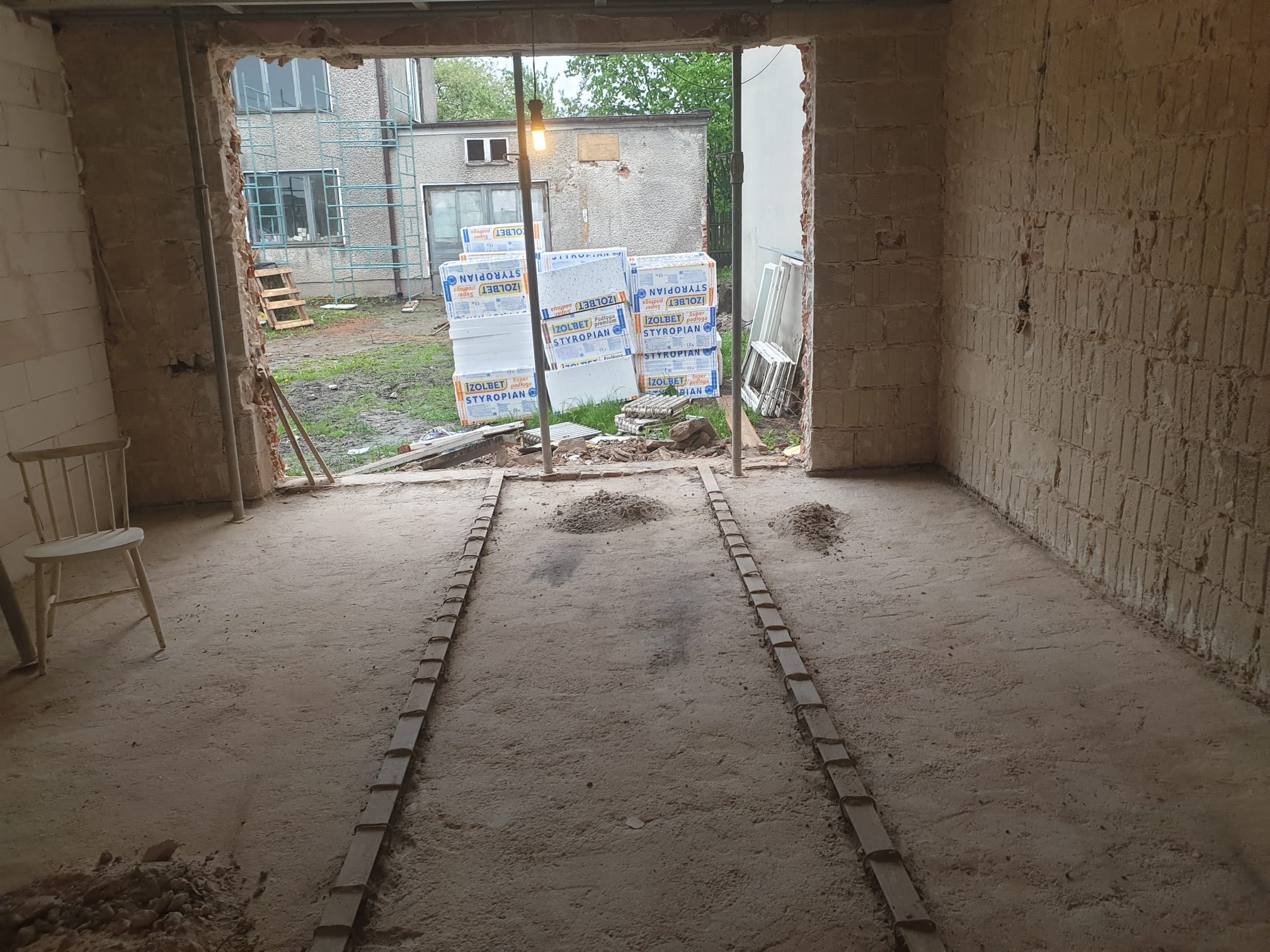 Wnętrze w trakcie prac przygotowawczych do wylewki, widoczne oznaczone linie poziomów, ściany w stanie surowym, na zewnątrz budynku składowane płyty styropianowe.