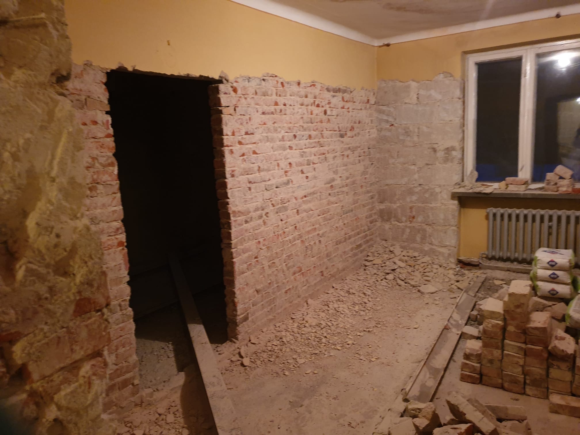 Pokój w trakcie rozbiórki: odsłonięta cegła na ścianach, gruz na podłodze, przygotowane worki z zaprawą, widoczny otwór drzwiowy w stanie surowym.