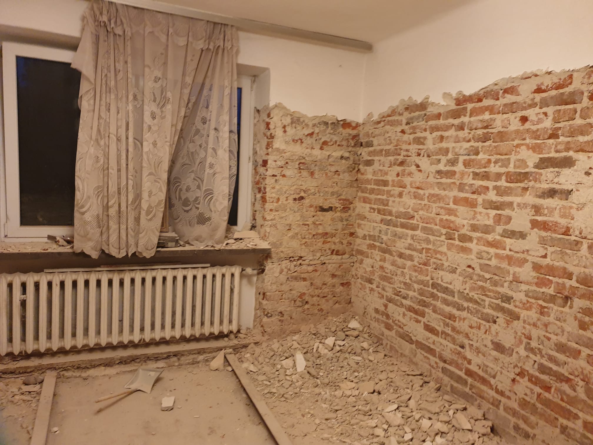 Odsłonięta ceglana ściana w trakcie prac remontowych, widoczne resztki tynku, grzejnik pod oknem z firankami i gruz na podłodze.