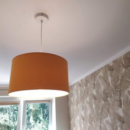 Adam Przybylski - Zawieszona, włączona lampa z abażurem w kolorze musztardowym, widok z dołu, z oknem i tapetą w liście w tle.