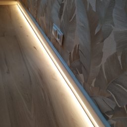 Adam Przybylski - Listwa przypodłogowa z wbudowanym oświetleniem LED, zamontowana przy ścianie z tapetą w motywy roślinne, obok gniazdko elektryczne.