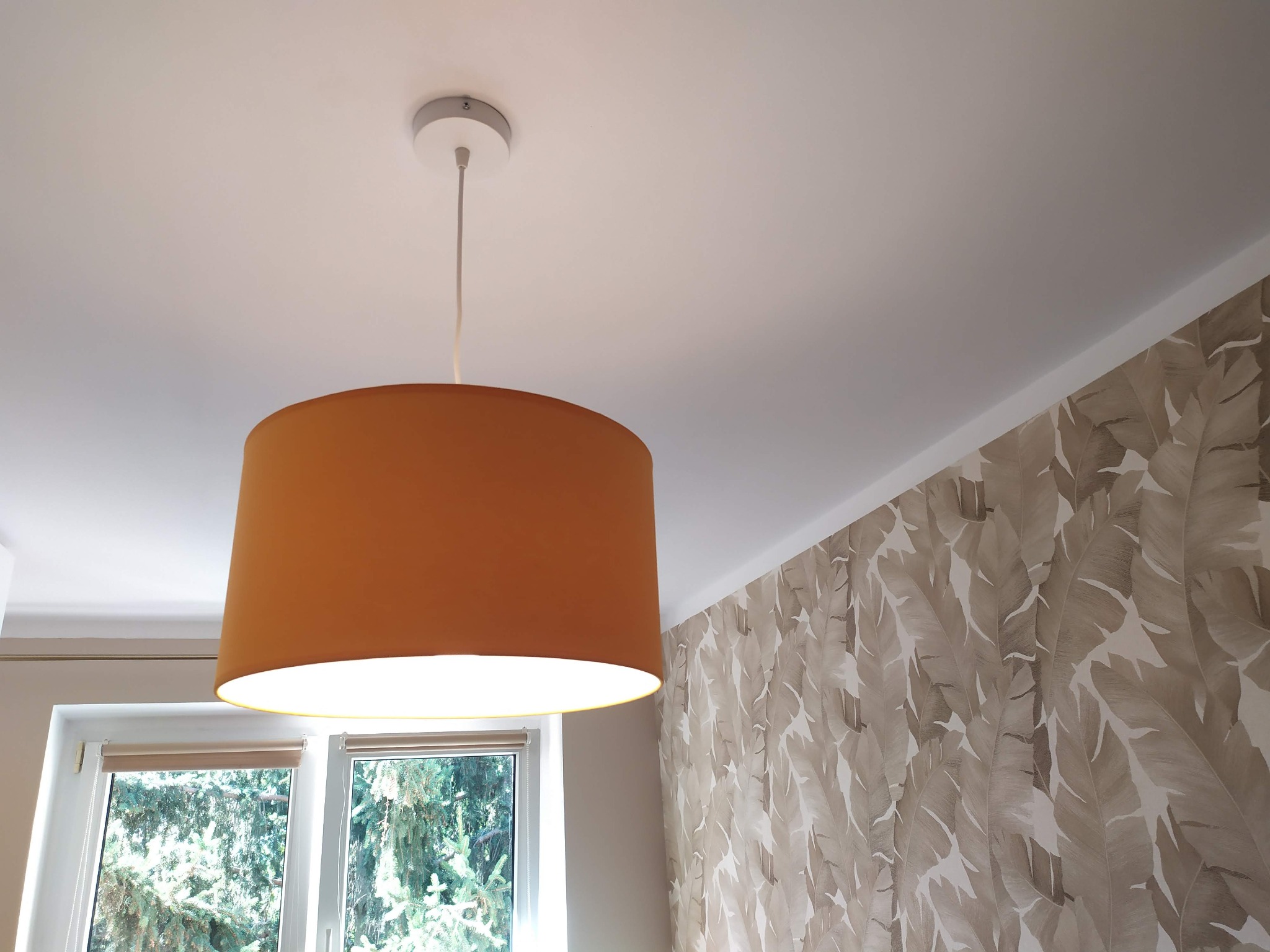 Zawieszona, włączona lampa z abażurem w kolorze musztardowym, widok z dołu, z oknem i tapetą w liście w tle.