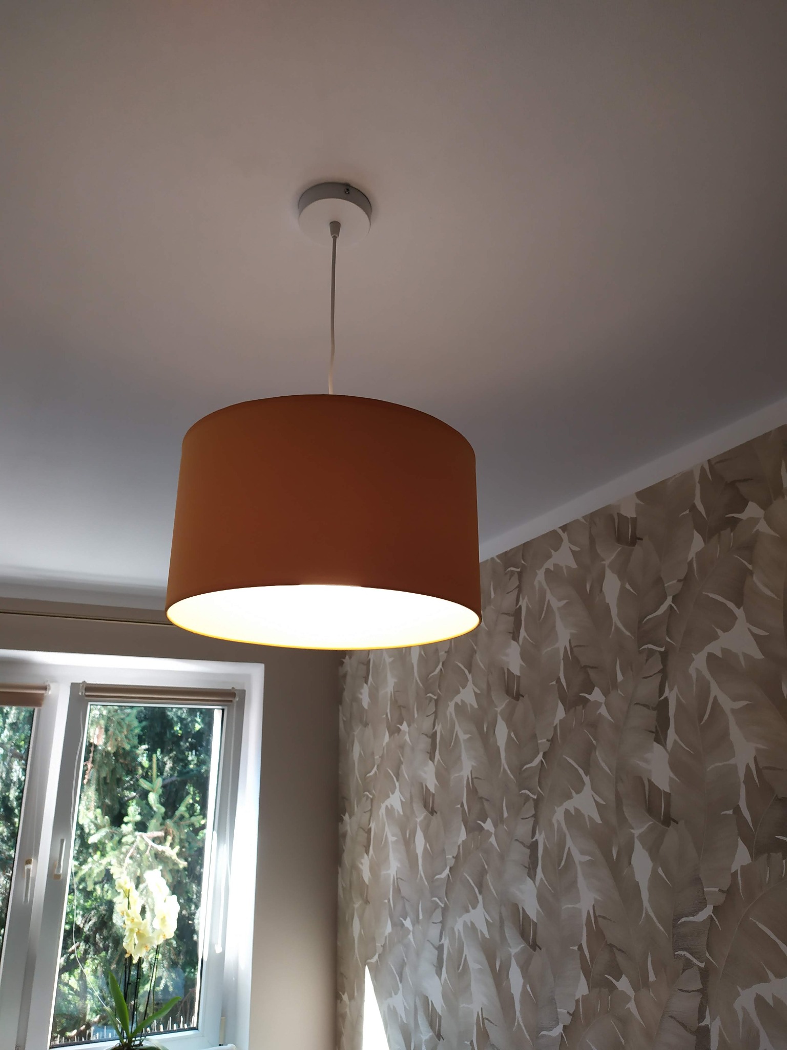 Brązowy abażur lampy wiszącej z włączonym światłem, widok z dołu na tle sufitu, okna z kwiatami i ściany w liście.