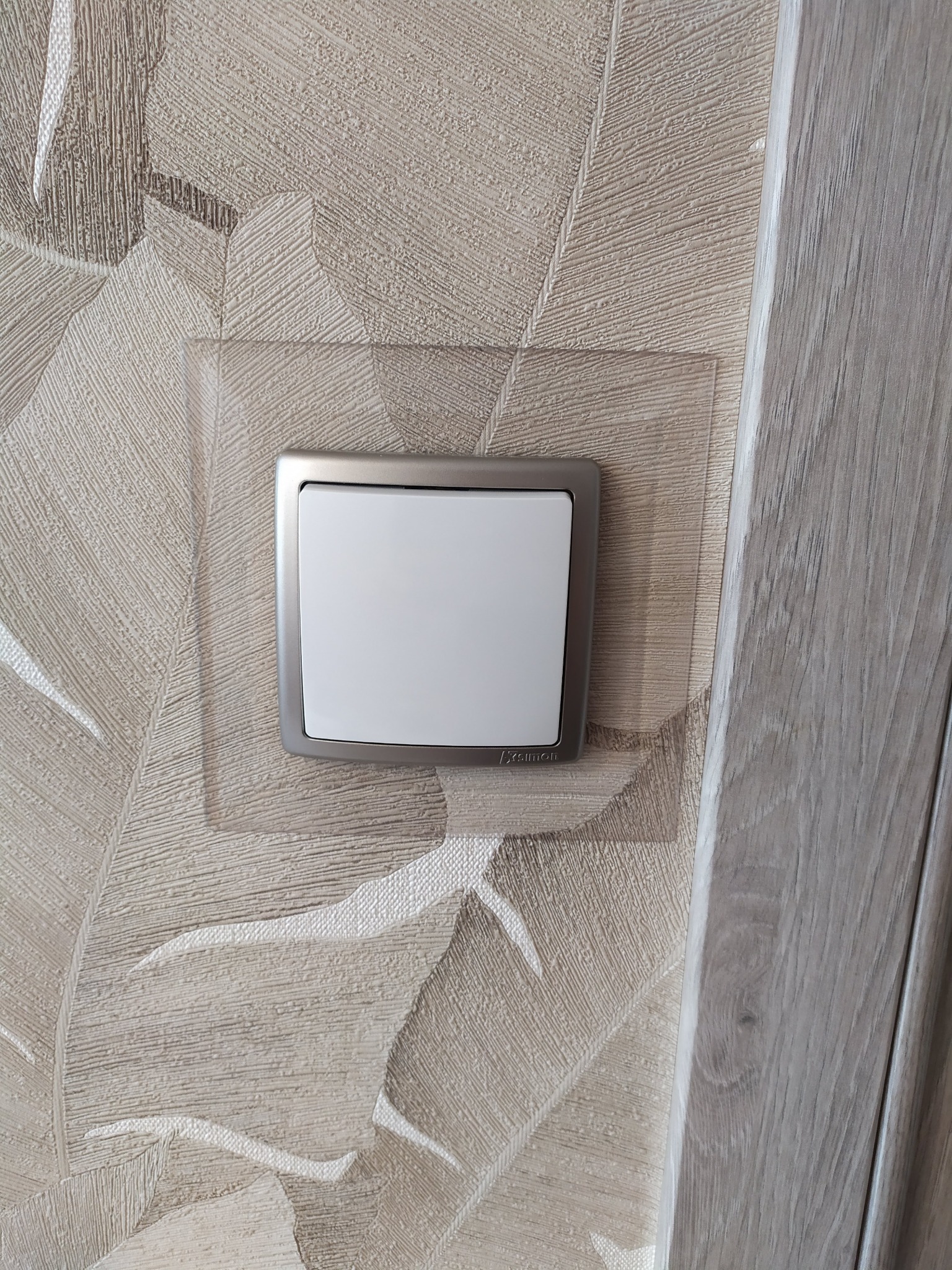 Biały, kwadratowy włącznik światła marki Simon z metalową ramką, zamontowany na beżowej ścianie z wzorem liści, obok drewnianej futryny drzwi.