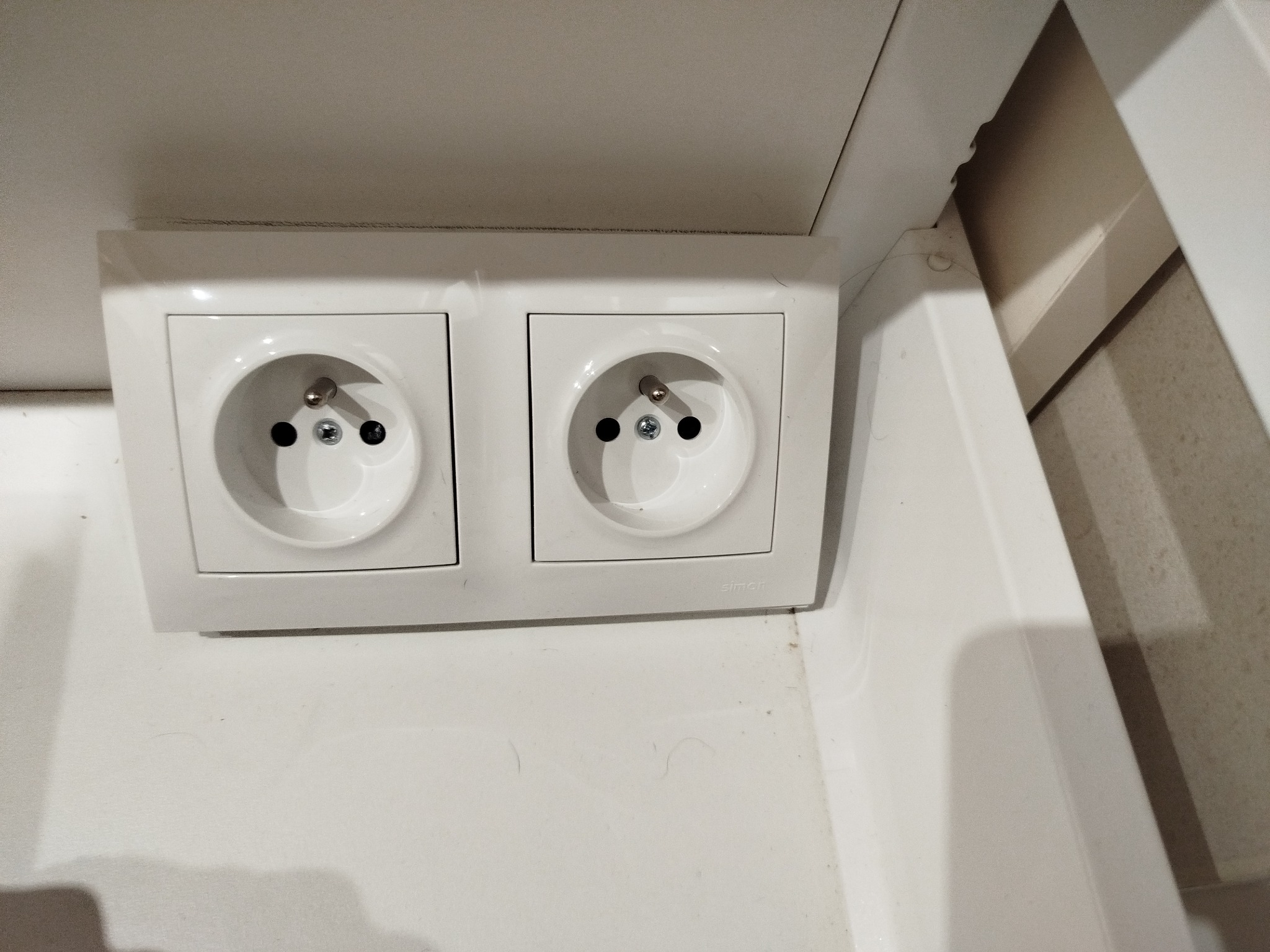 Dwa białe gniazdka elektryczne marki Simon zamontowane na białej ścianie, widoczne bolce uziemiające.