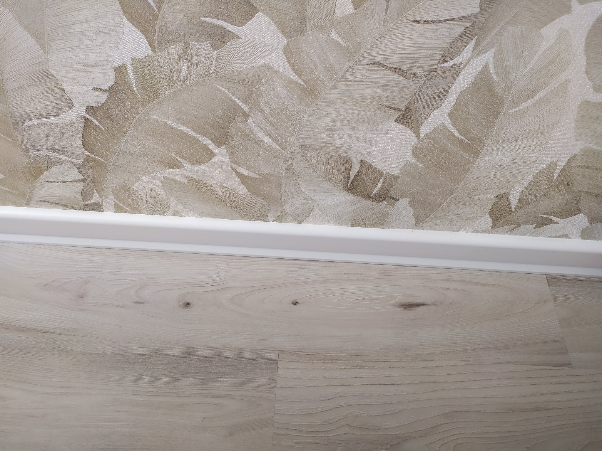 Fragment ściany z tapetą w liście i podłogi z paneli z białą listwą przypodłogową.
