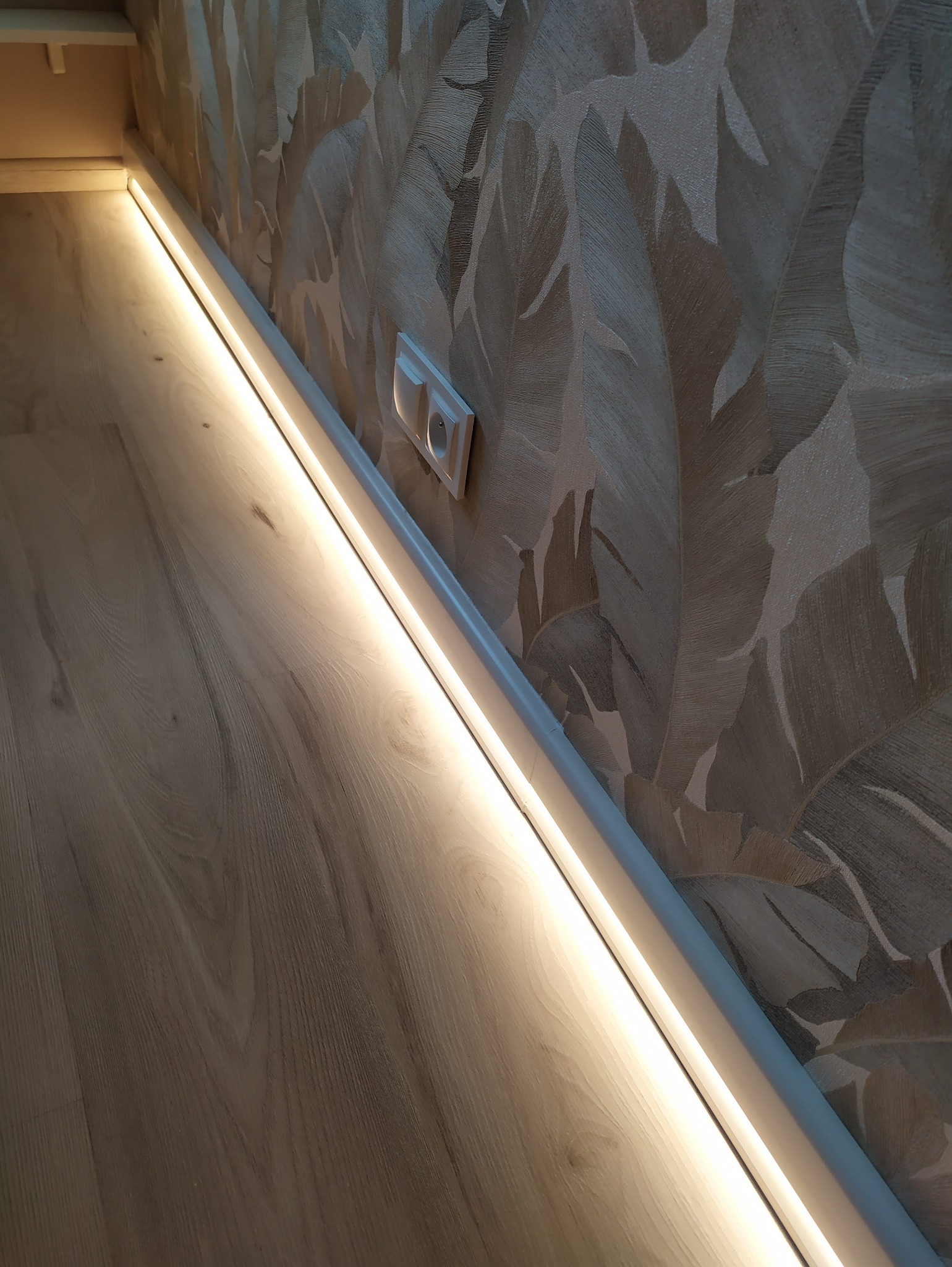Listwa przypodłogowa z wbudowanym oświetleniem LED, zamontowana przy ścianie z tapetą w motywy roślinne, obok gniazdko elektryczne.