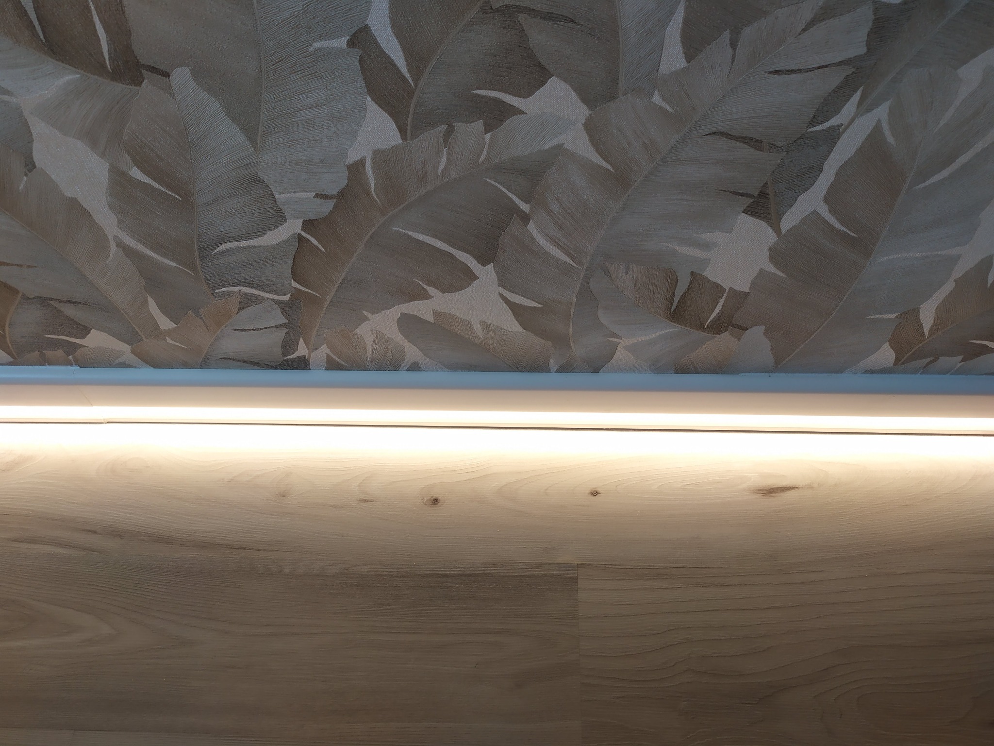 Ukryte oświetlenie LED w listwie przysufitowej, oświetlające drewnopodobną okładzinę ściany i tapetę w motywy roślinne.