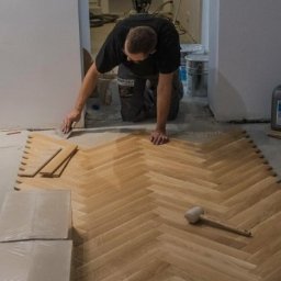 REMO DESIGN - Układanie parkietu w jodełkę: glazurnik docina deski przy ścianie, widoczne narzędzia, paczki desek, klej i śmieci budowlane.