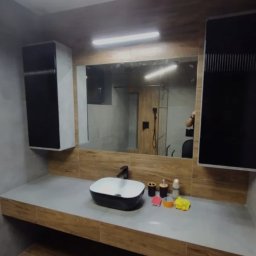 REMO DESIGN - Nowoczesna łazienka z umywalką nablatową w czarnej obudowie, drewnopodobnym blatem i szafkami wiszącymi w kolorze czarno-białym, widok na kabinę prysznicową w lustrze.