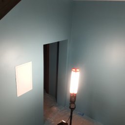 REMO DESIGN - Świeżo pomalowane na niebiesko ściany w pomieszczeniu z widocznym wejściem do innego pokoju, oświetlone lampą roboczą, z klapką rewizyjną na ścianie.