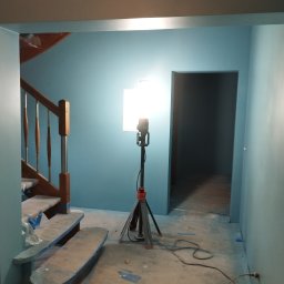 REMO DESIGN - Świeżo pomalowane na niebiesko ściany w Krosno Odrzańskie, widoczne schody zabezpieczone folią malarską oraz lampa budowlana na trójnogu oświetlająca wnętrze.