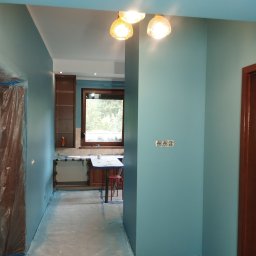 REMO DESIGN - Turkusowe ściany po malowaniu w trakcie remontu, zabezpieczone folią drzwi, widoczne gniazdka elektryczne i fragment kuchni w tle.