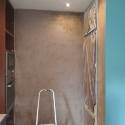 REMO DESIGN - Ściana w trakcie wykańczania z nałożoną gładzią w odcieniach beżu, zabezpieczone folią meble i drabina aluminiowa w pomieszczeniu z oświetleniem punktowym.